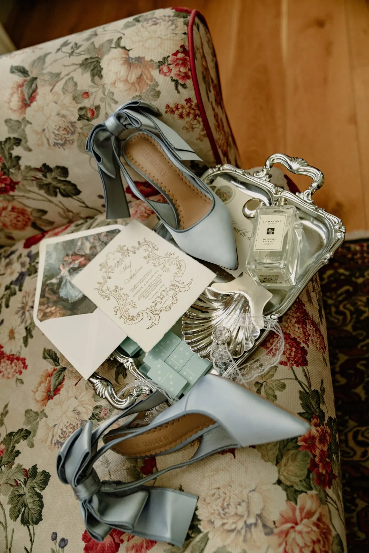 Pantofi de mireasă, invitație și parfum pe o canapea florală